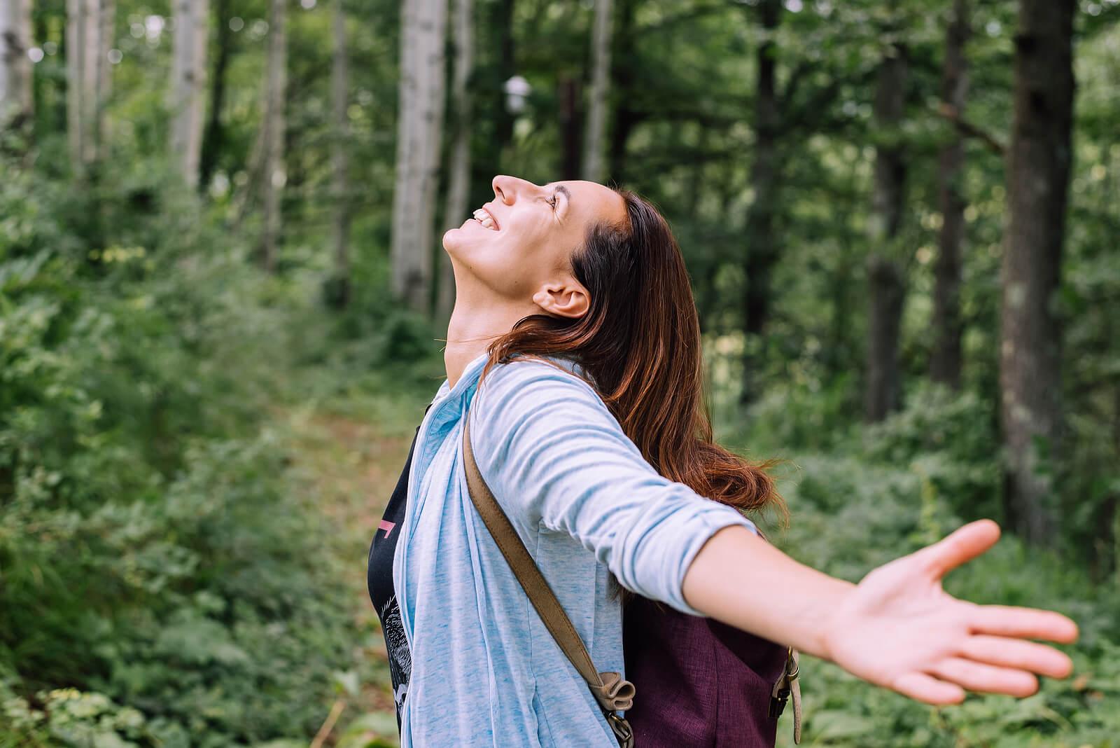 Image of a smiling woman standing in the middle of a forest looking up at the sky with her arms spread out. Overcome difficult emotions and begin healing with Adult IOP in Houston, TX.