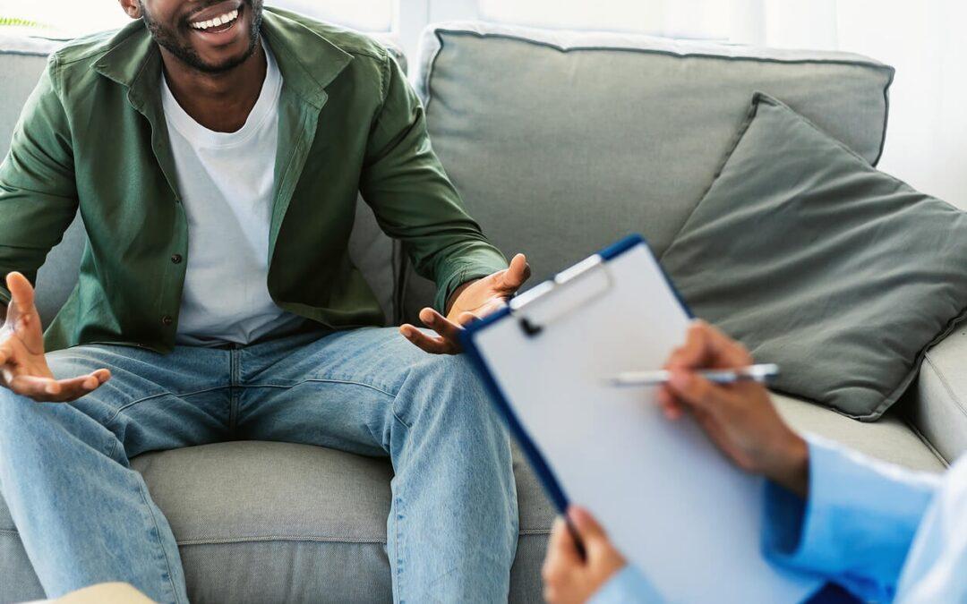 Image of a smiling man sitting on a couch speaking to a therapist. Overcome your anxiety, depression, and more with the support of adult intensive outpatient program in Houston, TX.