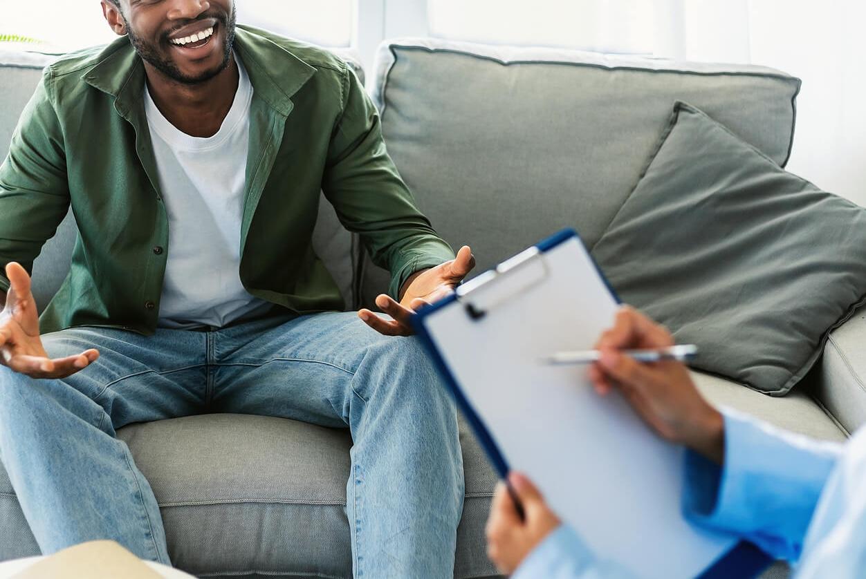 Image of a smiling man sitting on a couch speaking to a therapist. Overcome your anxiety, depression, and more with the support of adult intensive outpatient program in Houston, TX.