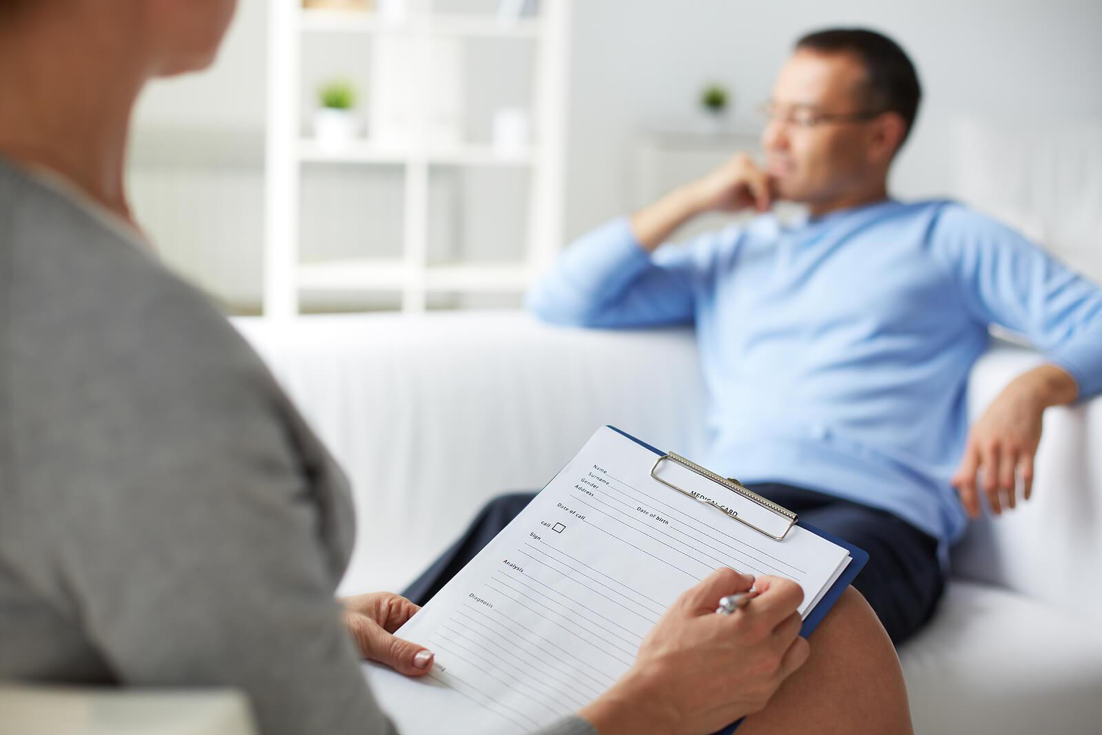 Image of a smiling man sitting on a couch while speaking to a woman holding a clipboard. Start healing from your depression in effective ways tailored to you with the support of Adult Intensive Outpatient Program in Houston, TX.