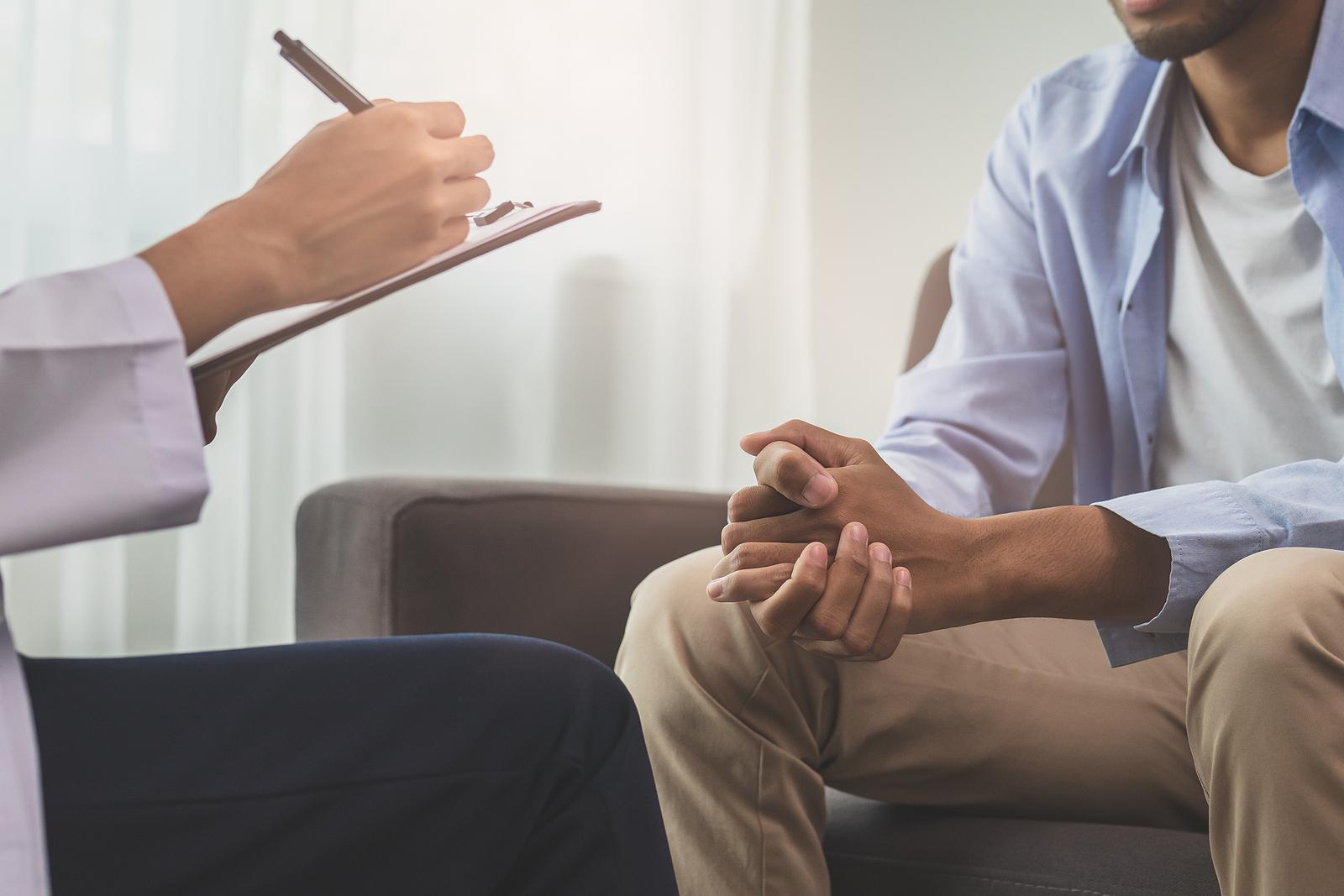 Image of a man sitting on a couch clasping his hands together while a woman writes on a clipboard opposite of him. If you're looking to build emotional resilience and improve your quality of life, find support with Adult Intensive Outpatient Program in Houston, TX.