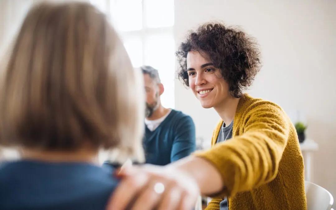 Image of people in group therapy with a woman smiling and resting her hand on another woman's shoulder showing support. With adult IOP in Houston, TX you can begin to cope in positive ways and find the support you need to begin healing from anxiety, depression, BPD, and more.