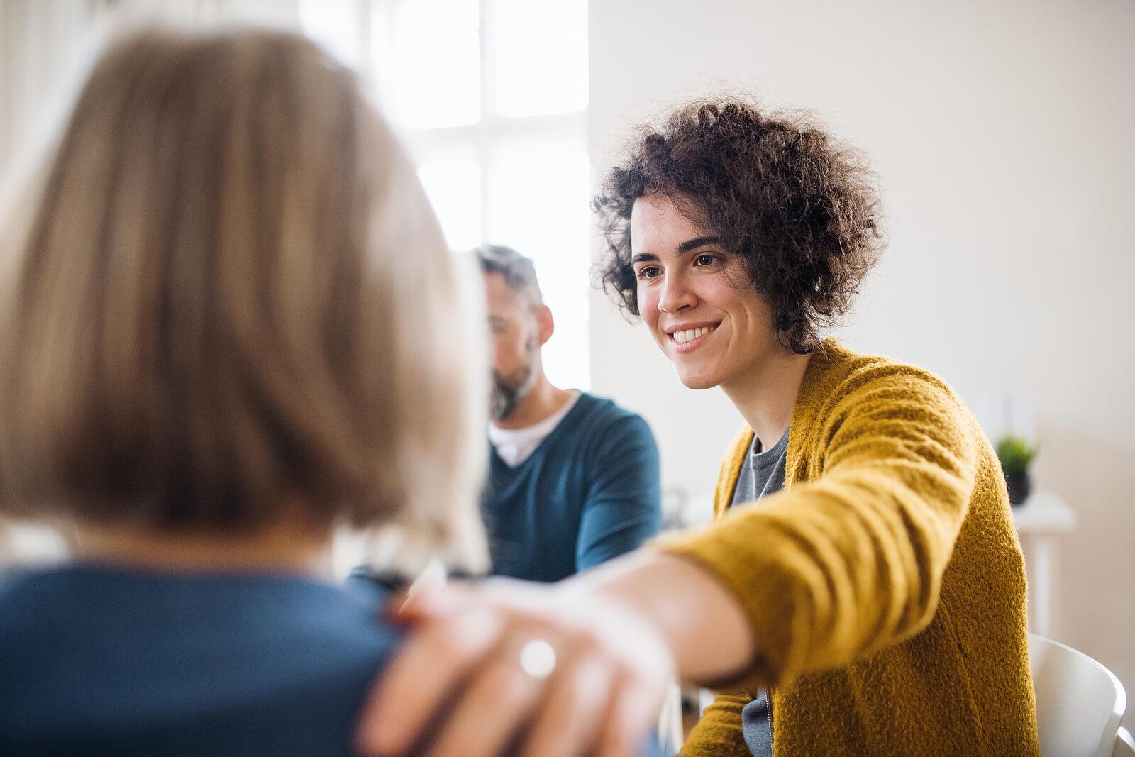 Image of people in group therapy with a woman smiling and resting her hand on another woman's shoulder showing support. With adult IOP in Houston, TX you can begin to cope in positive ways and find the support you need to begin healing from anxiety, depression, BPD, and more.