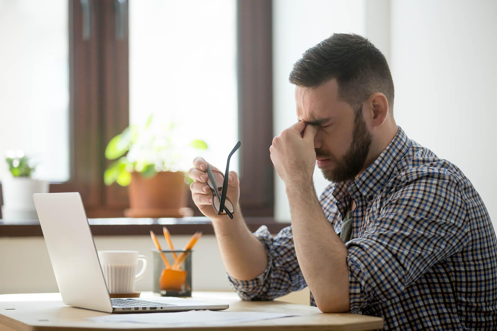 Stressed man sitting at a desk in front of a laptop closing his eyes and holding the bridge of his nose. If you struggle with being perfect all the time, find support from a compassionate therapist on healing with perfectionism treatment in Houston, TX.