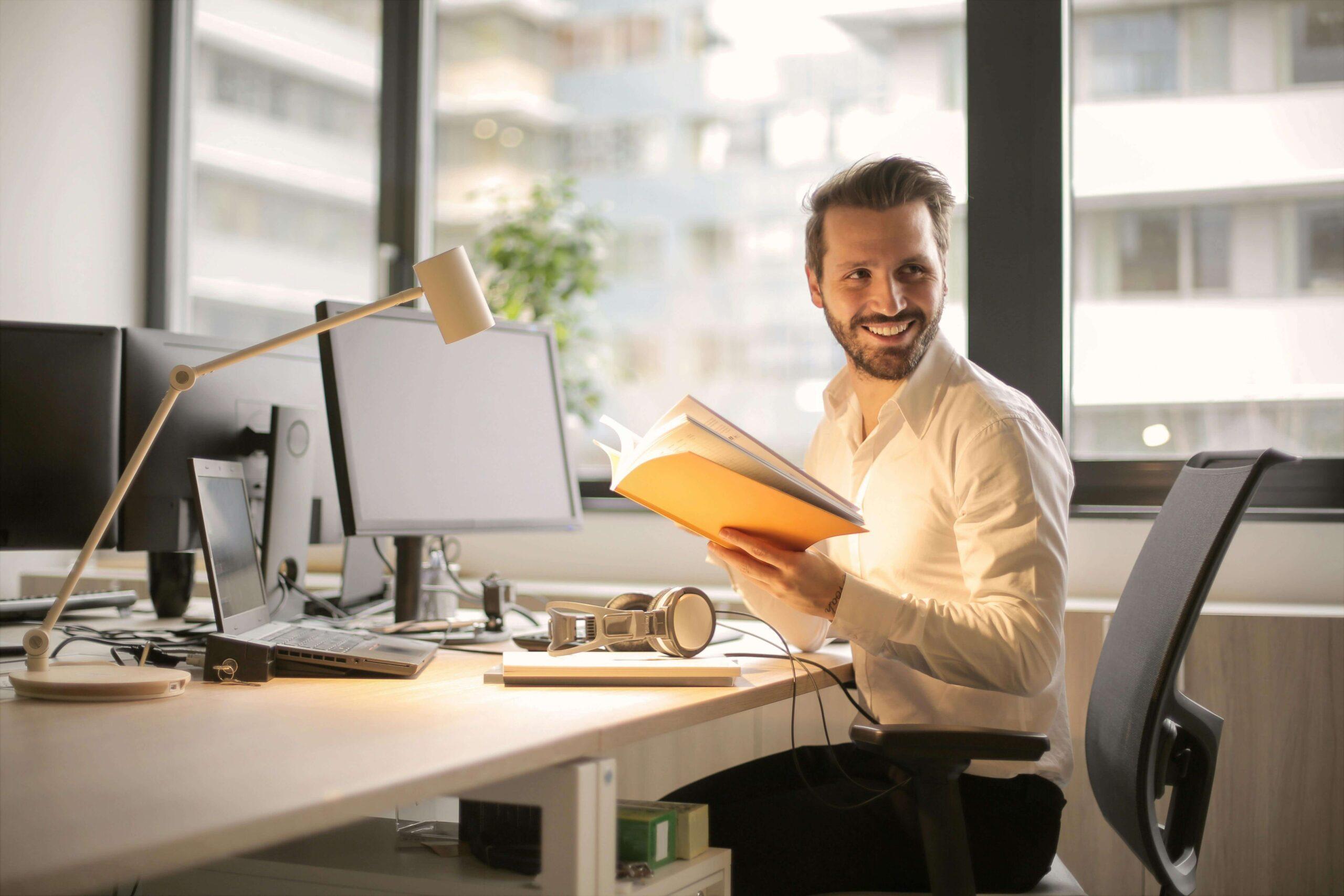 Happy professional man sitting at a office desk smiling and holding a book. With perfectionism treatment in Houston, TX you can learn to find self-compassion and openness to imperfection.