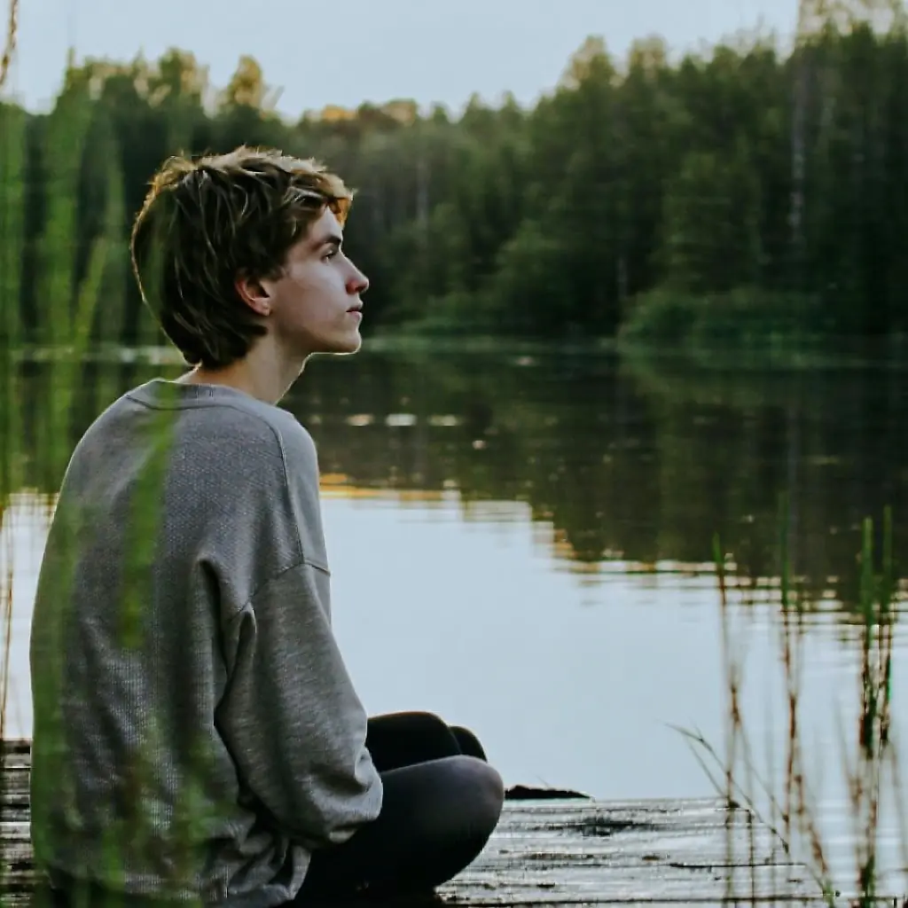 Teen sitting by a lake with trees all around looking thoughtful
