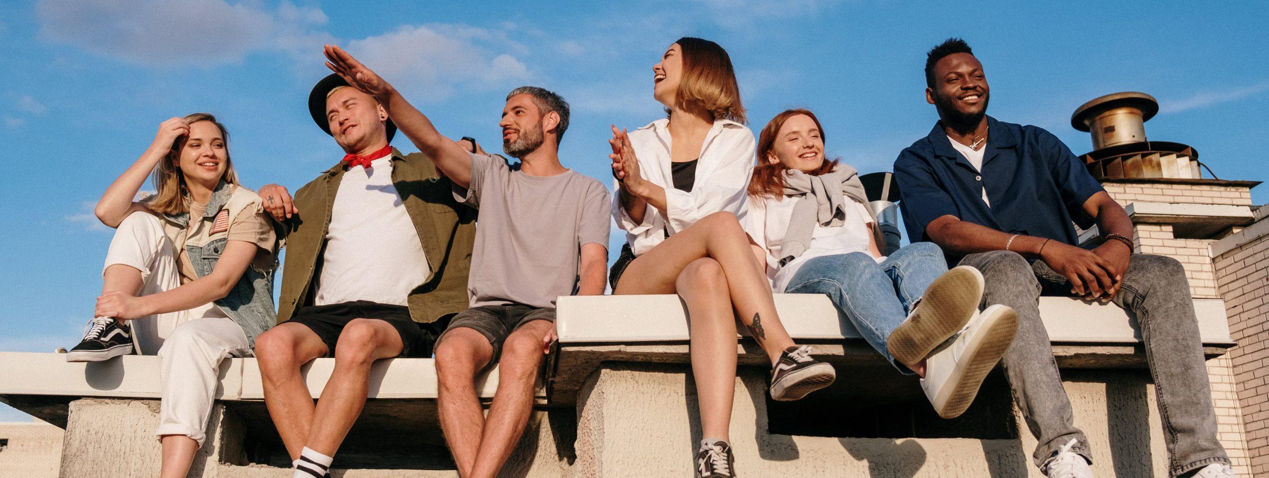 group of young people both males and females outside with a blue sky sitton on a sand colored bench. they are in various poses